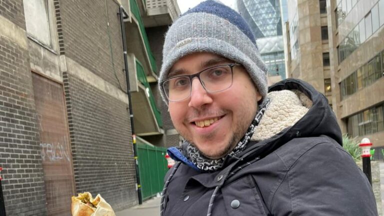 Ian smiling with a vegan falafel wrap from Eye Falafel in his hands, with The Shard in the background, wearing a winter hat and black coat