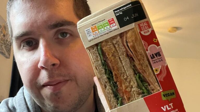 Ian holding a La Vie vegan VLT sandwich in its wrapper with a neutral expression